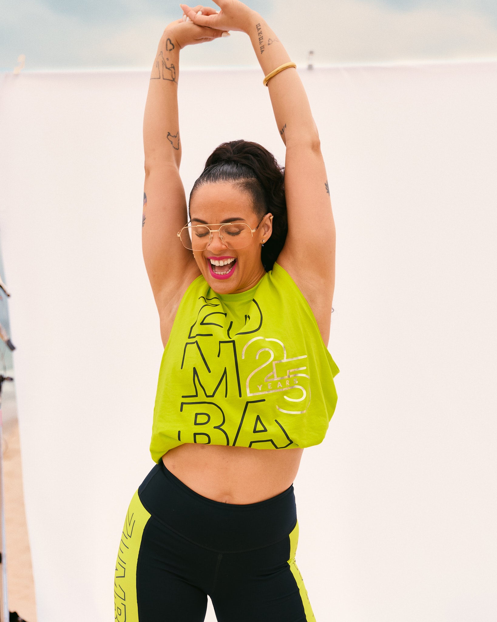 Neon yellow Zumba 25 Years cropped tank and black high-waisted leggings with neon yellow side panels. Female model wears the sleeveless cropped tank with fitted leggings, stretching with both arms raised in front of a white backdrop.