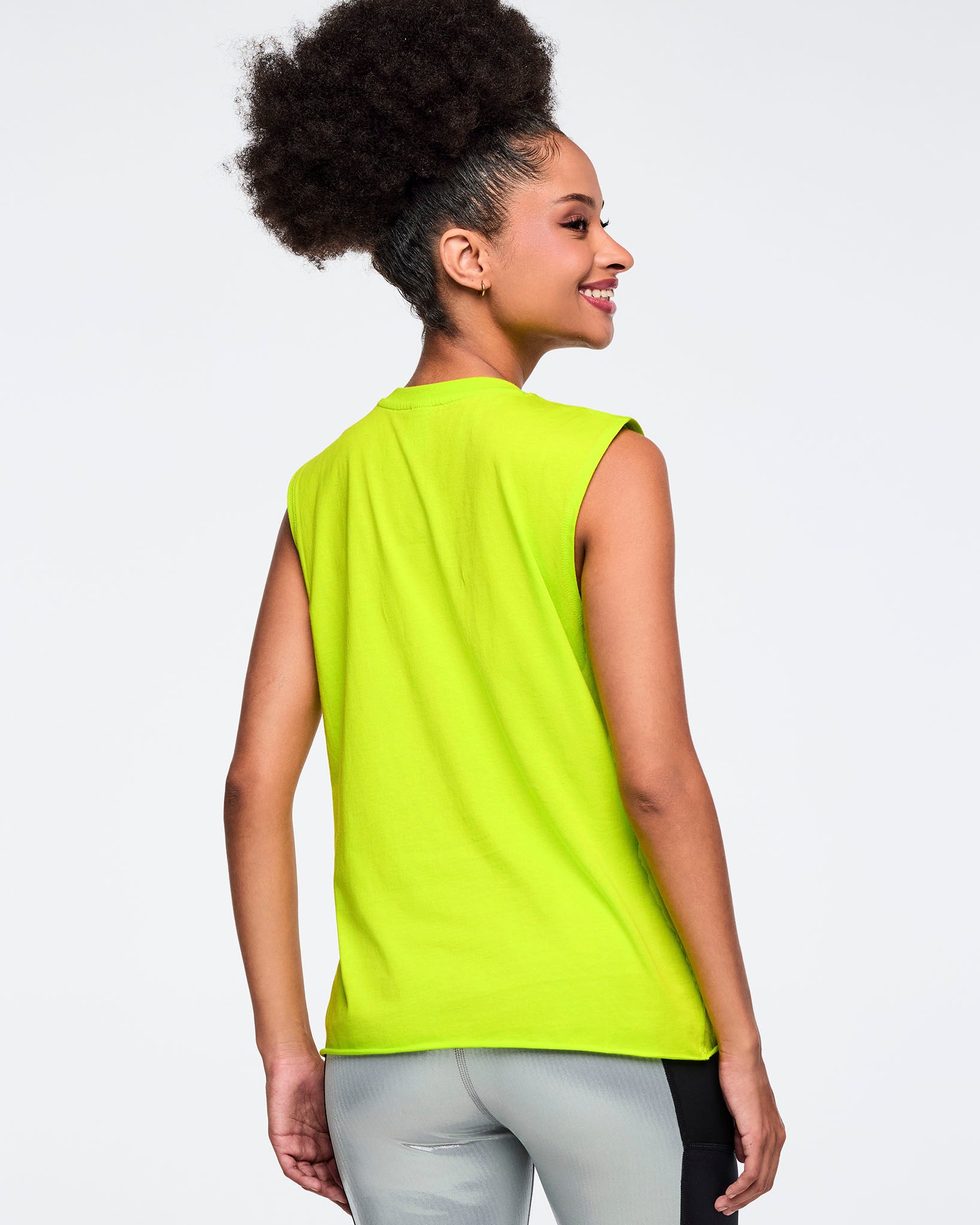 Neon green sleeveless Zumba tank with a clean solid back. Female model wears the relaxed-fit muscle tank with metallic silver leggings featuring black side panels, shown from the back against a light gray background.