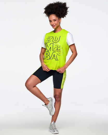Neon green sleeveless Zumba tank with oversized black logo graphics and silver “25 Years” detail. Female model wears the bright muscle tank layered over a white short-sleeve tee, black biker shorts with a neon green side panel and vertical Zumba logo, and silver athletic sneakers, posing against a light gray background.