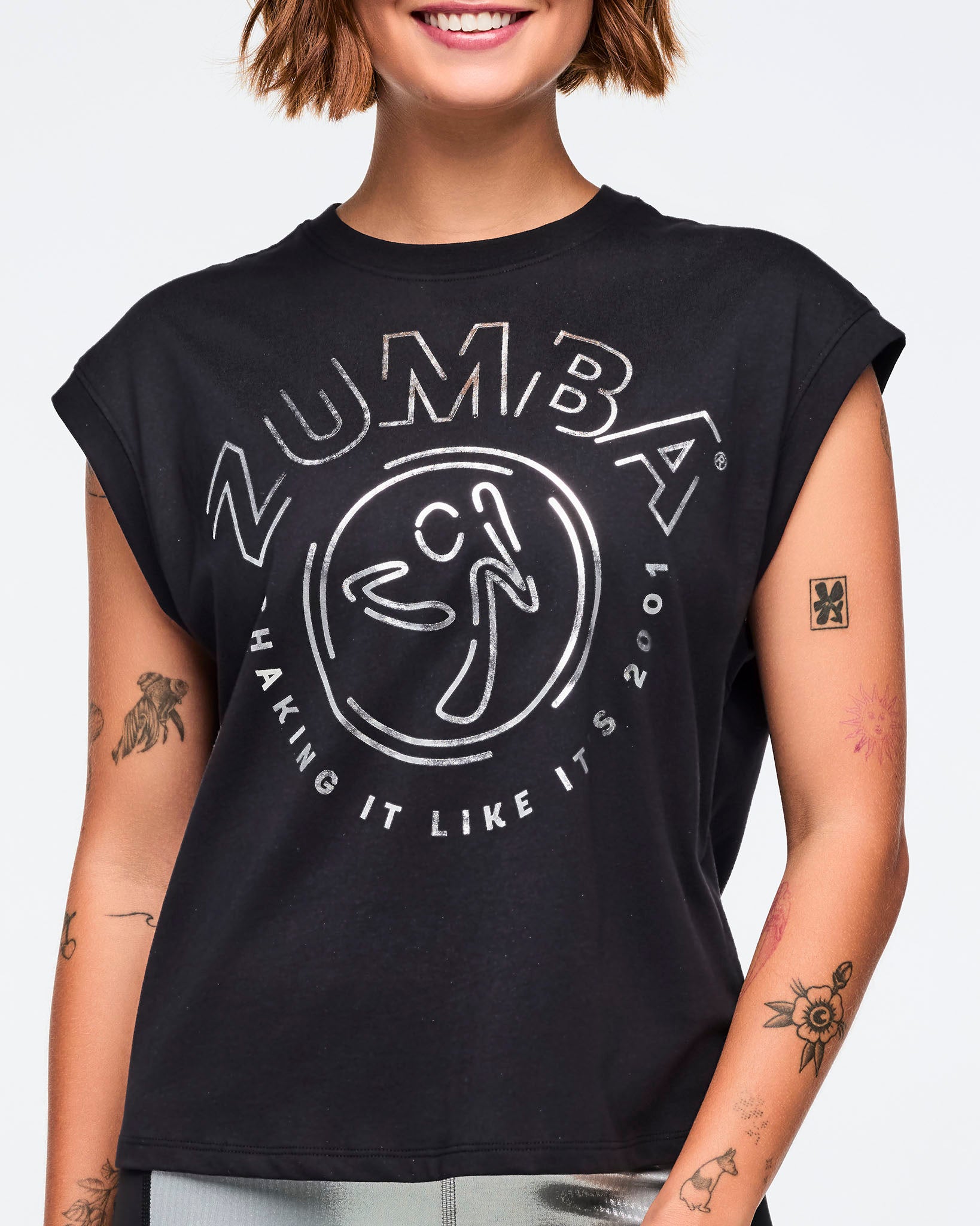 Black cap-sleeve Zumba graphic top with a bold silver front logo and slogan. Female model wears the relaxed-fit top with metallic silver leggings featuring black side panels, shown in a close front view against a light gray background.