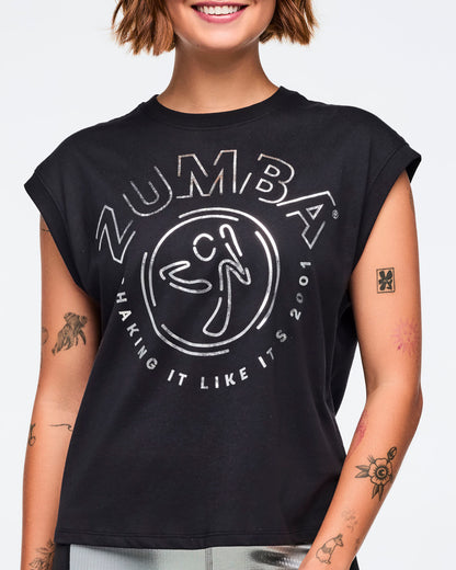 Black cap-sleeve Zumba graphic top with a bold silver front logo and slogan. Female model wears the relaxed-fit top with metallic silver leggings featuring black side panels, shown in a close front view against a light gray background.
