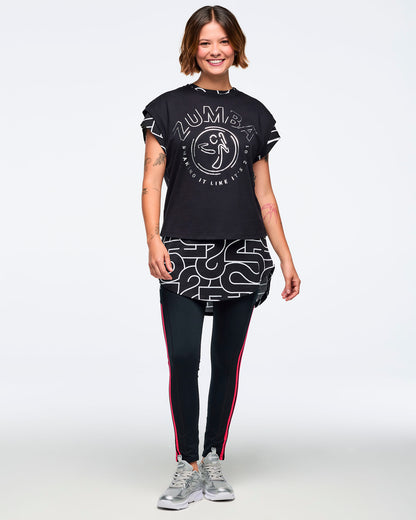 Black cap-sleeve Zumba graphic top with a large silver front logo. Female model wears the top layered over a long black-and-silver patterned shirt, black leggings with bright red side stripes, and silver athletic sneakers, standing in a full-body front view against a light gray background.