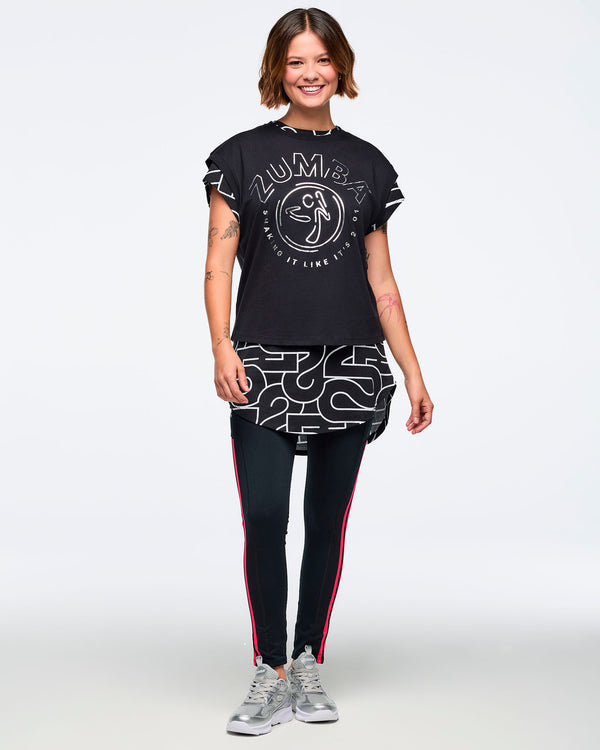 Black cap-sleeve Zumba graphic top with a large silver front logo. Female model wears the top layered over a long black-and-silver patterned shirt, black leggings with bright red side stripes, and silver athletic sneakers, standing in a full-body front view against a light gray background.