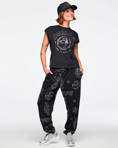 Black cap-sleeve Zumba graphic top with a large silver front logo. Female model wears the relaxed-fit top with black jogger pants covered in silver Zumba prints, a black baseball cap, and silver athletic sneakers, posing against a light gray background.