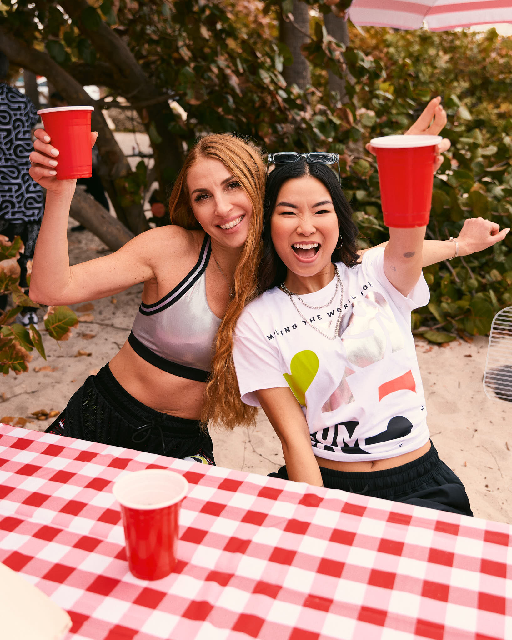 Two female models smile at a picnic table; one wears the silver sports bra with black joggers, and the other wears the loose white cropped Zumba tee with black joggers.