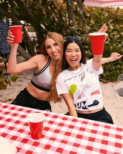 Two female models smile at a picnic table; one wears the silver sports bra with black joggers, and the other wears the loose white cropped Zumba tee with black joggers.