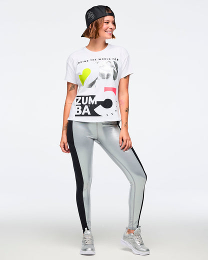 White short-sleeve Zumba graphic T-shirt with oversized black, silver, neon yellow, and pink anniversary artwork on the front. Female model wears the relaxed-fit tee with metallic silver leggings featuring black side panels, silver athletic sneakers, and a black baseball cap worn backward, shown in a full-body front view against a light gray background.