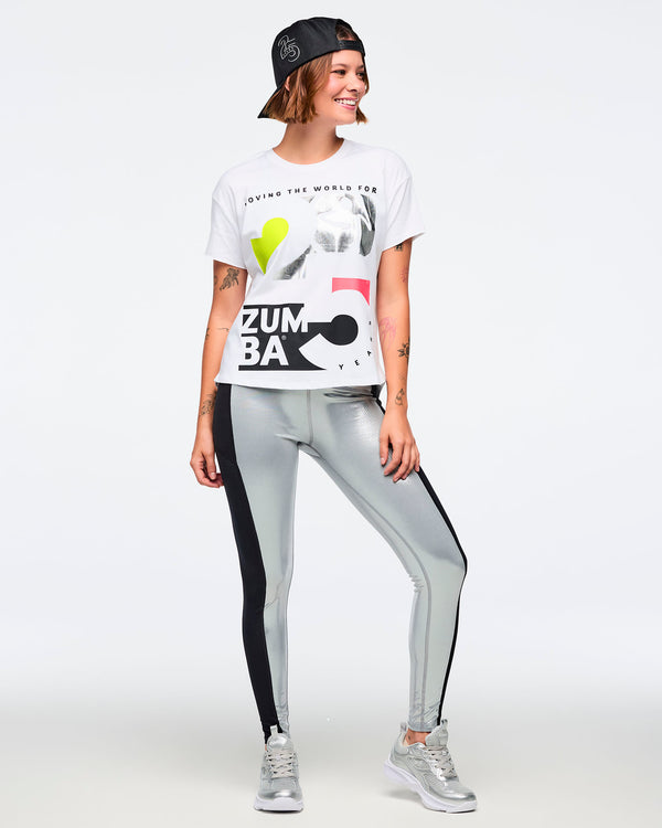 White short-sleeve Zumba graphic T-shirt with oversized black, silver, neon yellow, and pink anniversary artwork on the front. Female model wears the relaxed-fit tee with metallic silver leggings featuring black side panels, silver athletic sneakers, and a black baseball cap worn backward, shown in a full-body front view against a light gray background.