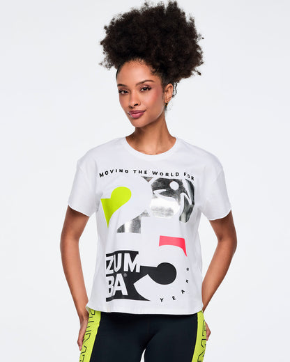 White short-sleeve Zumba graphic T-shirt with bold black, silver, neon yellow, and pink anniversary artwork and “Moving the World For 25” text on the front. Female model wears the relaxed-fit tee with black leggings featuring neon yellow side panels, shown in a front view against a light gray background.