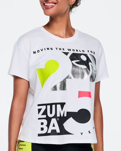 White short-sleeve Zumba graphic T-shirt with oversized black, silver, neon yellow, and pink 25th anniversary artwork on the front. Female model wears the relaxed-fit tee with black leggings featuring neon yellow side panels, shown in a close front view against a light gray background.