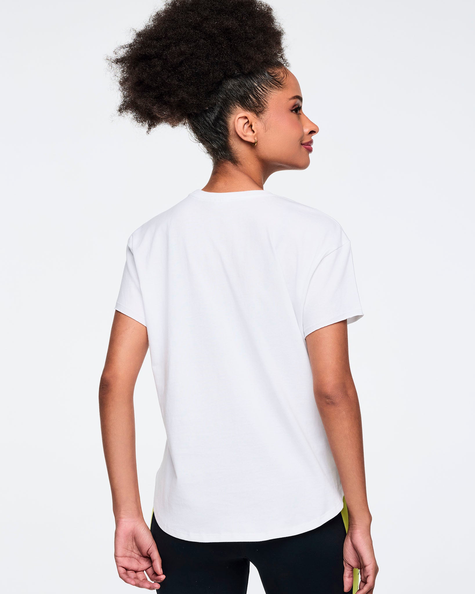 White short-sleeve Zumba graphic T-shirt with a plain solid back. Female model wears the relaxed-fit tee with black leggings featuring neon yellow side panels, shown from the back against a light gray background.