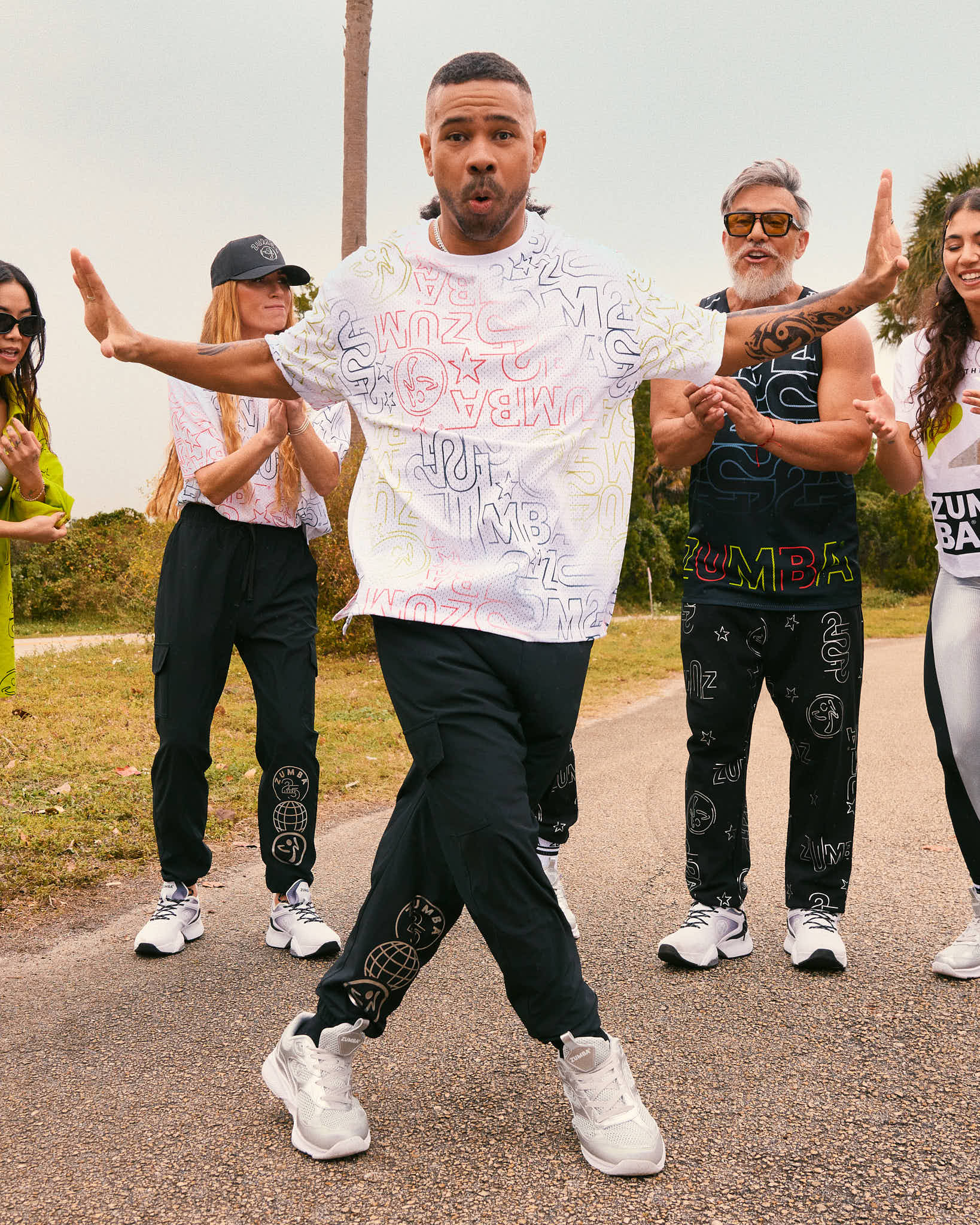 Male model wears the loose white allover-print tee with black joggers and silver athletic sneakers, dancing on a road while other models in coordinated Zumba activewear clap behind him.