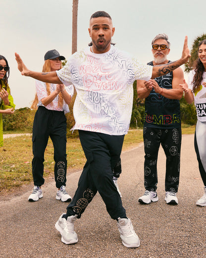 Male model wears the loose white allover-print tee with black joggers and silver athletic sneakers, dancing on a road while other models in coordinated Zumba activewear clap behind him.