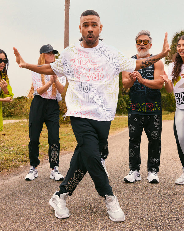 Male model wears the loose white allover-print tee with black joggers and silver athletic sneakers, dancing on a road while other models in coordinated Zumba activewear clap behind him.
