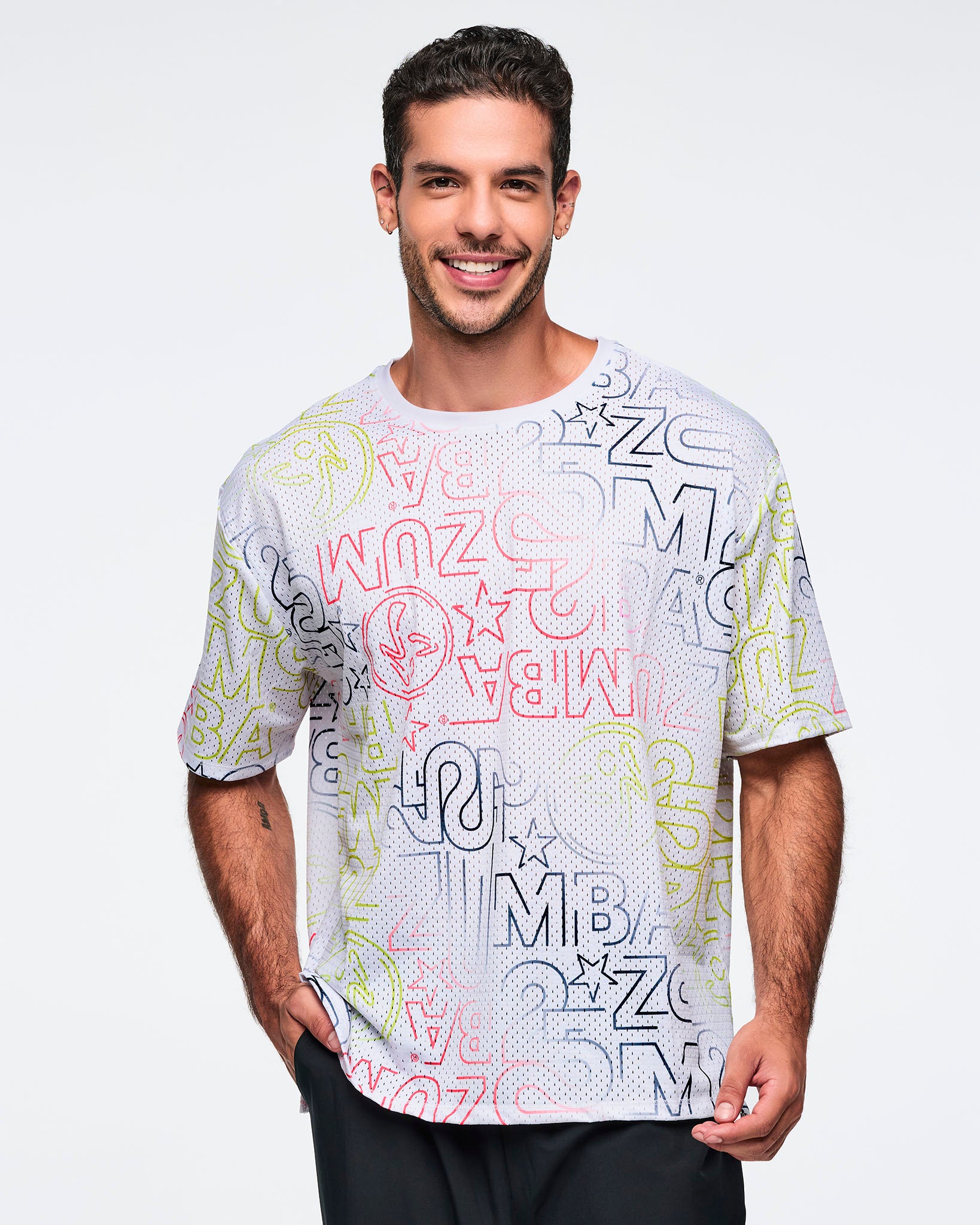 White oversized mesh Zumba top with multicolor allover logo print. Male model wears the relaxed short-sleeve top with black athletic pants, shown in a front view against a light gray background.