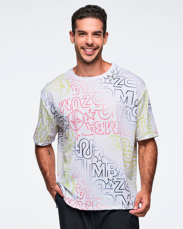 White oversized mesh Zumba top with multicolor allover logo print. Male model wears the relaxed short-sleeve top with black athletic pants, shown in a front view against a light gray background.
