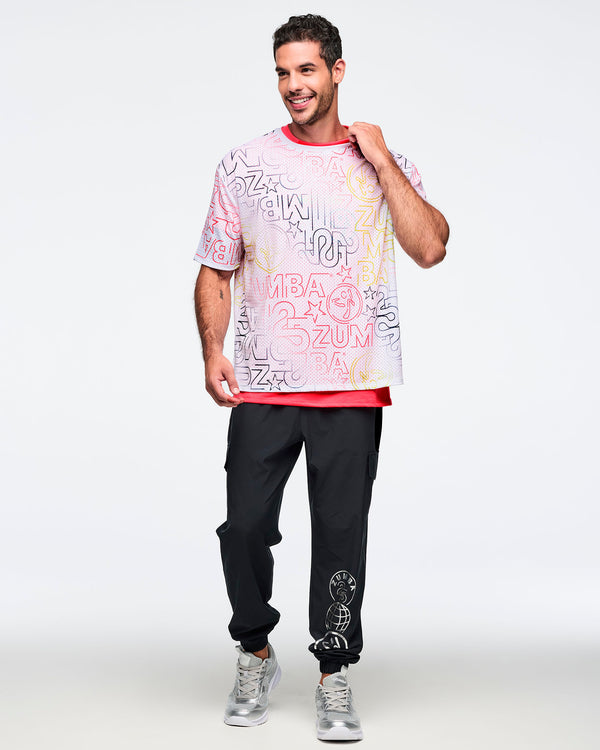 White oversized mesh Zumba top with multi-color all-over logo print layered over a bright coral pink T-shirt. Male model wears the relaxed short-sleeve top with black jogger pants and silver athletic sneakers, shown in a full-body front view against a light gray background.