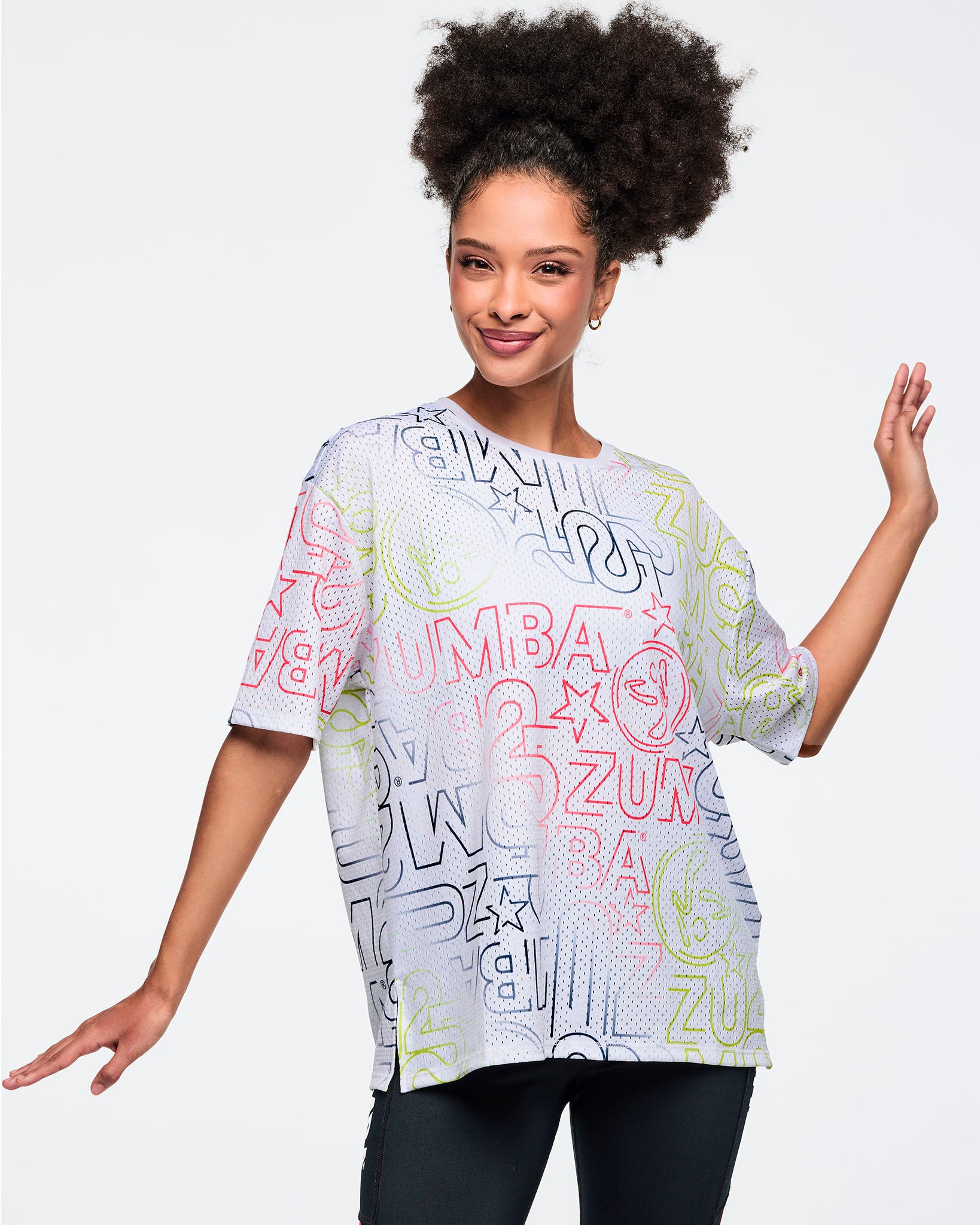 White oversized mesh Zumba top with multicolor allover logo print. Female model wears the relaxed short-sleeve top with black leggings, shown in a partial front view against a light gray background.