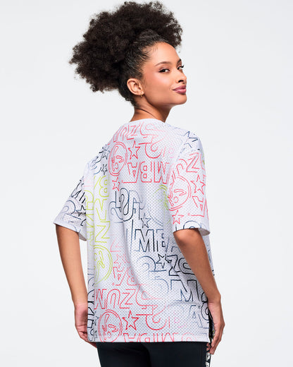 White oversized mesh Zumba top with multicolor allover logo print. Female model wears the relaxed short-sleeve top, shown in a close-up view highlighting the crew neckline and graphic details against a light gray background.