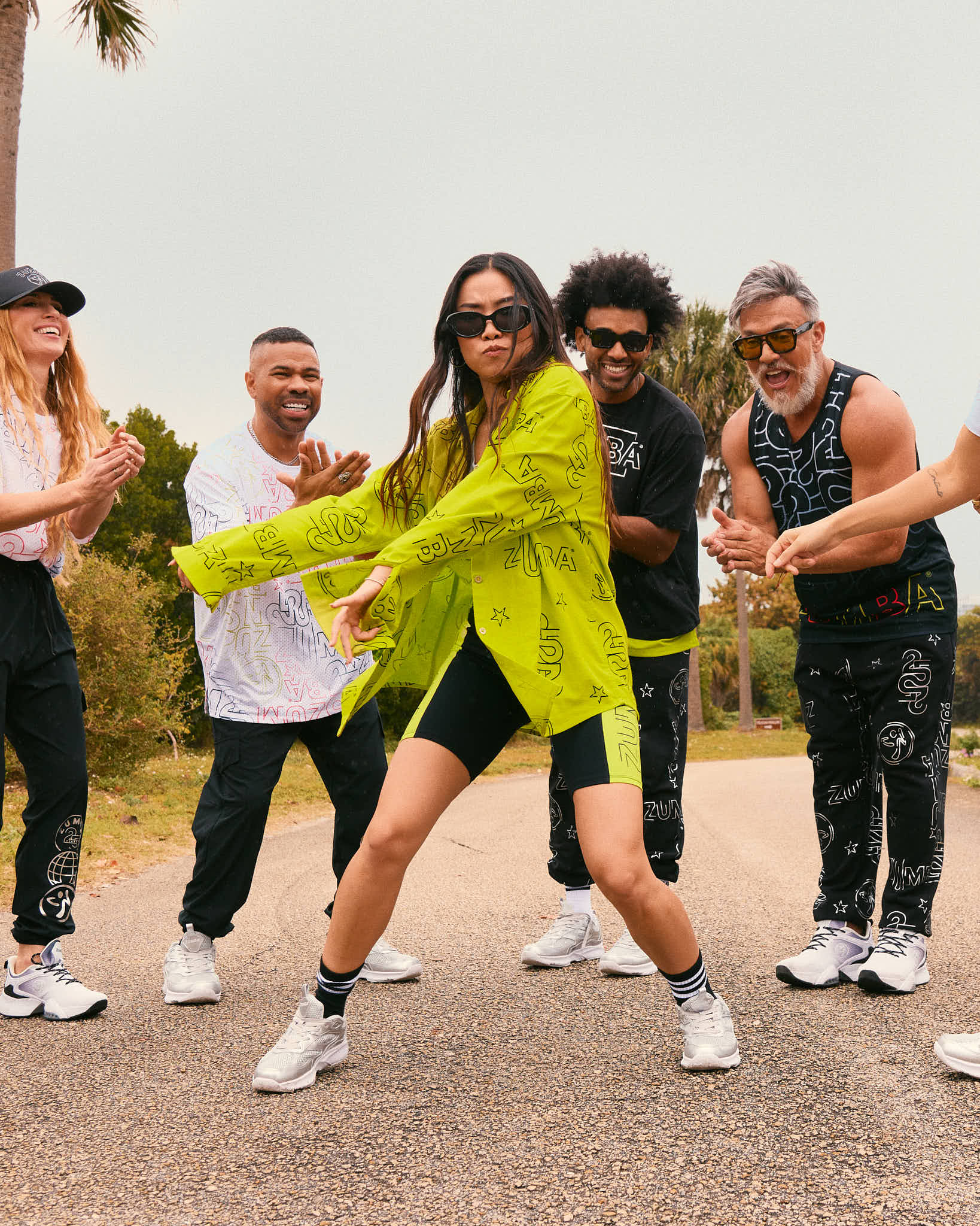 Female model wears the oversized shirt, fitted black shorts, black striped crew socks, silver sneakers, and black sunglasses while dancing on a road with other models in Zumba activewear.