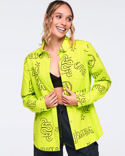Neon green long-sleeve button-down shirt with black allover Zumba logo print. Female model wears the relaxed collared shirt open over a black bra top with black cargo-style pants, shown in a front view against a light gray background.