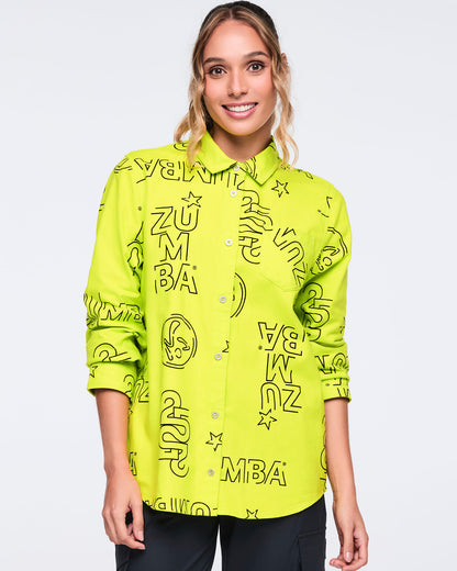 Neon green long-sleeve button-down shirt with black allover Zumba logo print. Female model wears the relaxed collared shirt buttoned up with black cargo-style pants, shown in a front view against a light gray background.