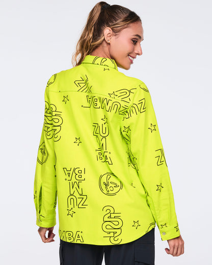 Neon green long-sleeve button-down shirt with black allover Zumba logo print. Female model wears the relaxed collared shirt with black cargo-style pants, shown from the back against a light gray background.
