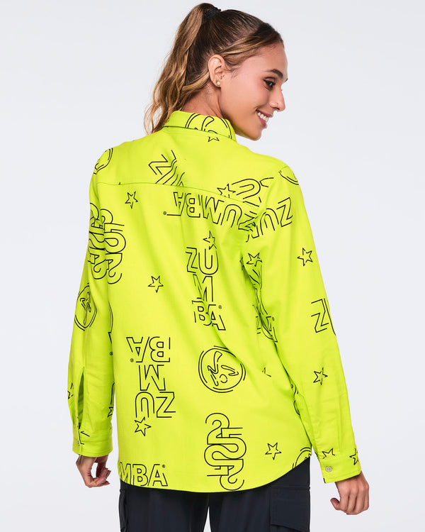 Neon green long-sleeve button-down shirt with black allover Zumba logo print. Female model wears the relaxed collared shirt with black cargo-style pants, shown from the back against a light gray background.