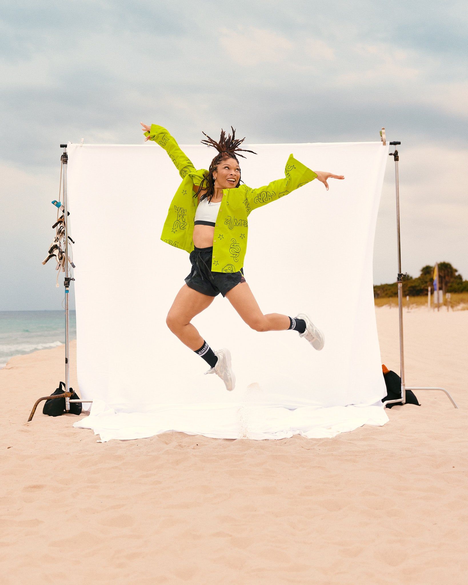 Female model jumps on the beach wearing the oversized long-sleeve shirt, black shorts, black crew socks, and silver sneakers in front of a white backdrop.