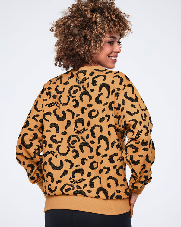 Back view of a tan crewneck sweatshirt with an allover black leopard print and ribbed cuffs, collar, and hem. Female model wears the long-sleeve sweatshirt with black fitted leggings, posing with one hand at her hip and looking over her shoulder against a light gray background.