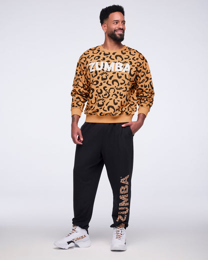 Tan long-sleeve crewneck sweatshirt with an allover black leopard print and large white Zumba logo across the chest. Male model wears the sweatshirt with black jogger sweatpants featuring a leopard-print Zumba graphic down one leg and white high-top sneakers, standing with one hand in his pocket against a light gray background.