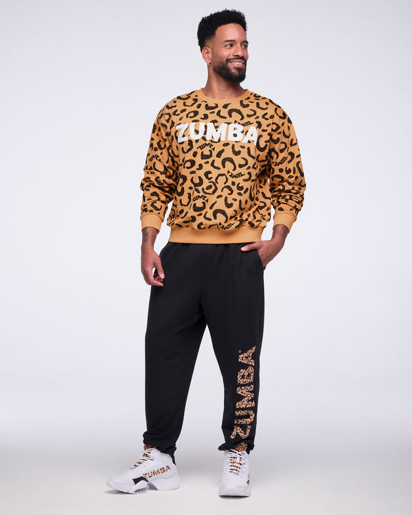 Tan long-sleeve crewneck sweatshirt with an allover black leopard print and large white Zumba logo across the chest. Male model wears the sweatshirt with black jogger sweatpants featuring a leopard-print Zumba graphic down one leg and white high-top sneakers, standing with one hand in his pocket against a light gray background.