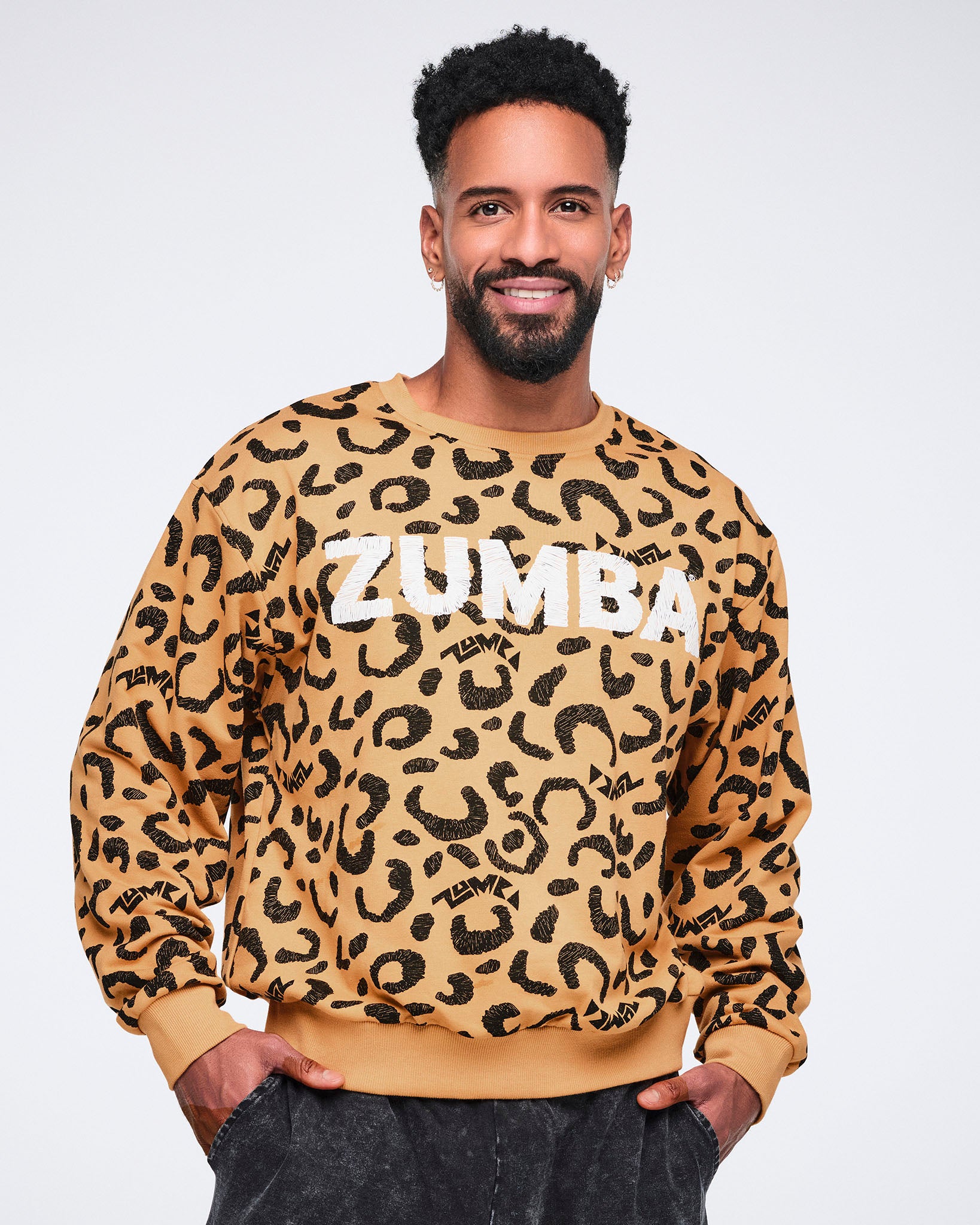 Tan leopard-print sweatshirt with black abstract spots, white “Zumba” lettering, and ribbed cuffs and hem, shown on a smiling male model wearing black mesh wide-leg pants with tan leopard-print side stripes.