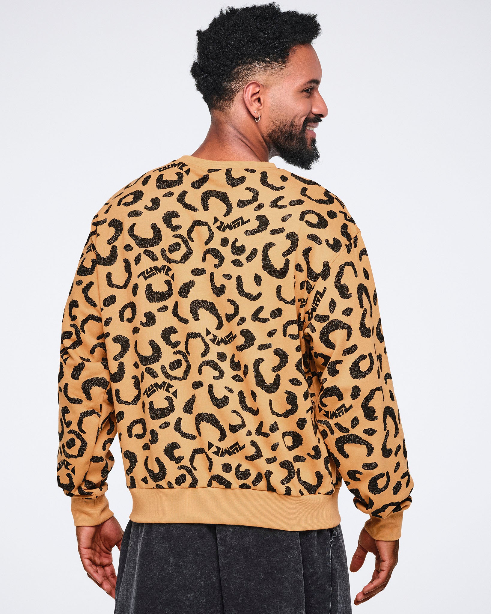 Back view of a tan crewneck sweatshirt with an allover black leopard print and ribbed cuffs, collar, and hem. Male model wears the long-sleeve sweatshirt with dark charcoal washed pants, standing turned slightly to the side and smiling against a light gray background.