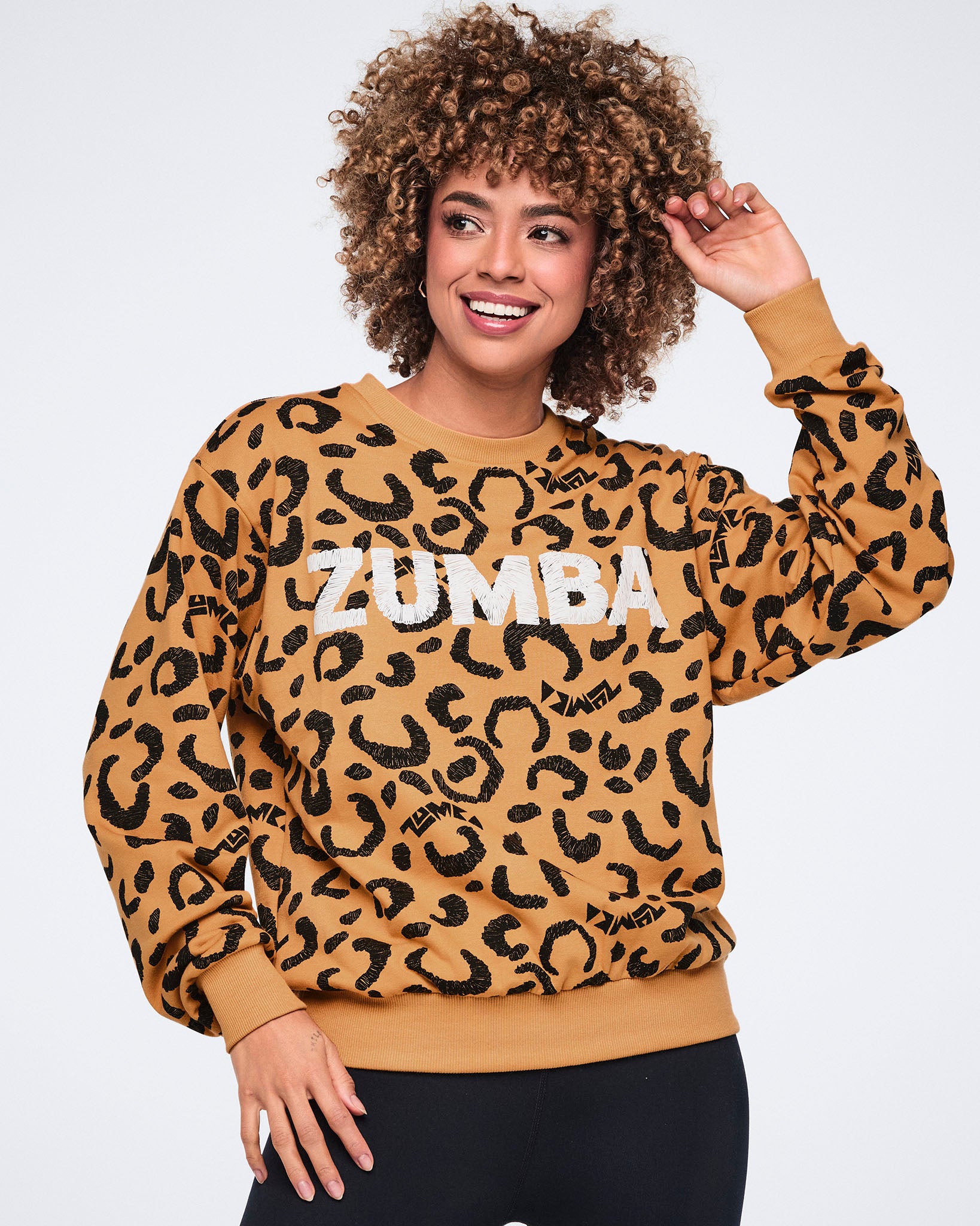 Tan long-sleeve crewneck sweatshirt with an allover black leopard print and large white Zumba logo across the chest. Female model wears the sweatshirt with black fitted leggings, smiling with one hand raised near her hair against a light gray background.