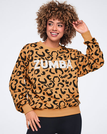 Tan long-sleeve crewneck sweatshirt with an allover black leopard print and large white Zumba logo across the chest. Female model wears the sweatshirt with black fitted leggings, smiling with one hand raised near her hair against a light gray background.