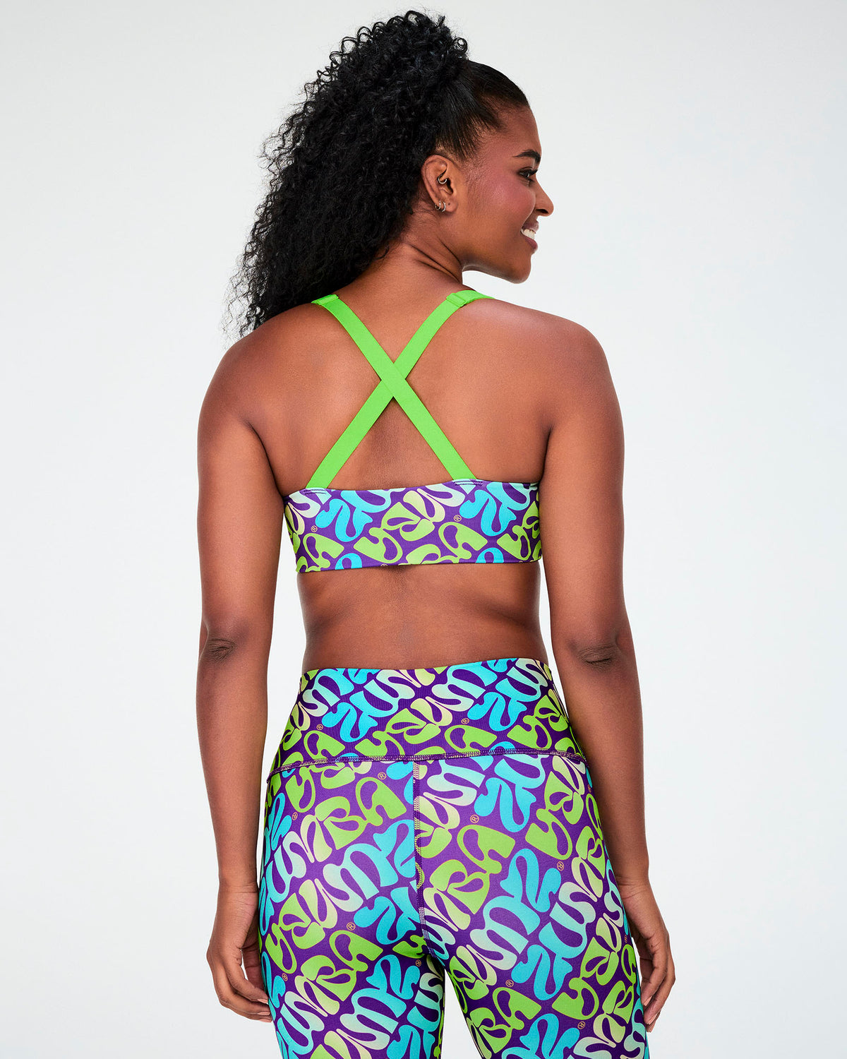 Shown from the back, the warped 'ZUMBA' print bra with criss-cross neon green straps, worn with matching print leggings.