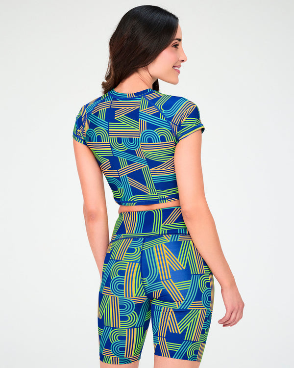 Back view of a female model in the blue retro striped-letter crop top and matching biker shorts, the green, light blue, and orange print continuing across the back.