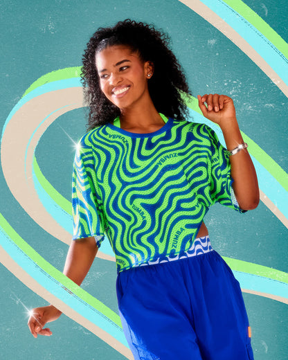 Female model in a neon green and royal blue wavy print mesh boxy crop top with curving 'ZUMBA' text and a blue neckband, paired with blue cargo pants, against an aqua background with swirling stripes.