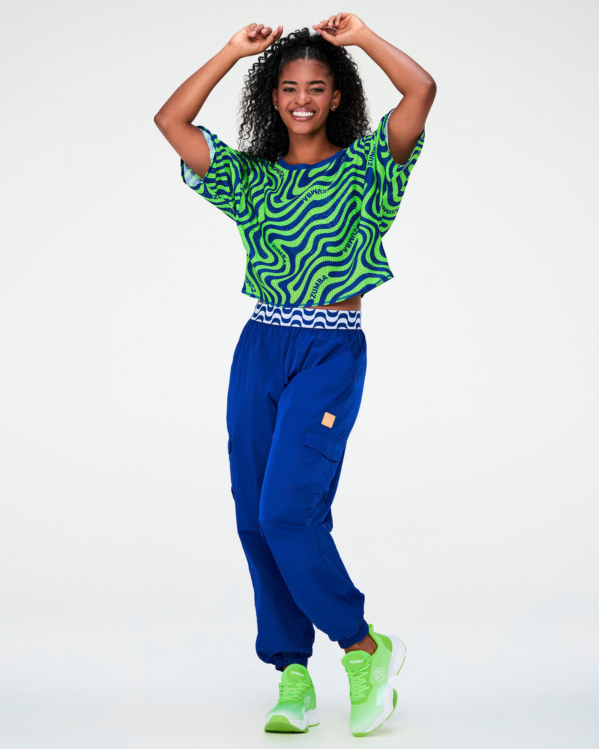 Full view of a female model with arms raised in the neon green and blue wavy mesh crop top, paired with blue parachute cargo pants with a blue and white waistband and neon green shoes.