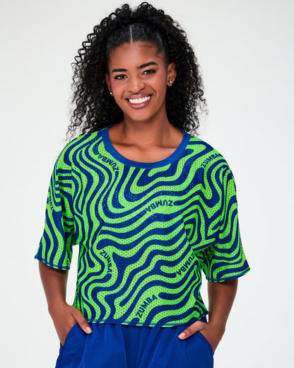 Female model in the neon green and blue wavy print mesh boxy crop top, its wide elbow-length sleeves and boxy cropped hem shown over blue cargo pants.