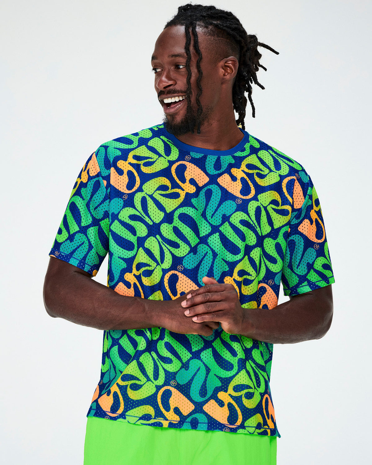 Male model in a blue mesh boxy top with an all-over diagonal print of warped 'ZUMBA' lettering in neon green, aqua, and orange, small Zumbito logos, and a blue neckband, paired with neon green pants.