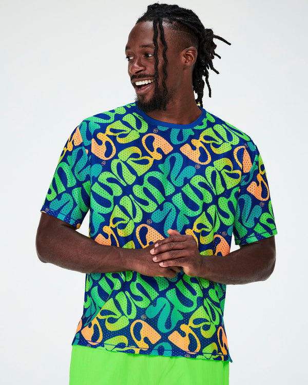 Male model in a blue mesh boxy top with an all-over diagonal print of warped 'ZUMBA' lettering in neon green, aqua, and orange, small Zumbito logos, and a blue neckband, paired with neon green pants.