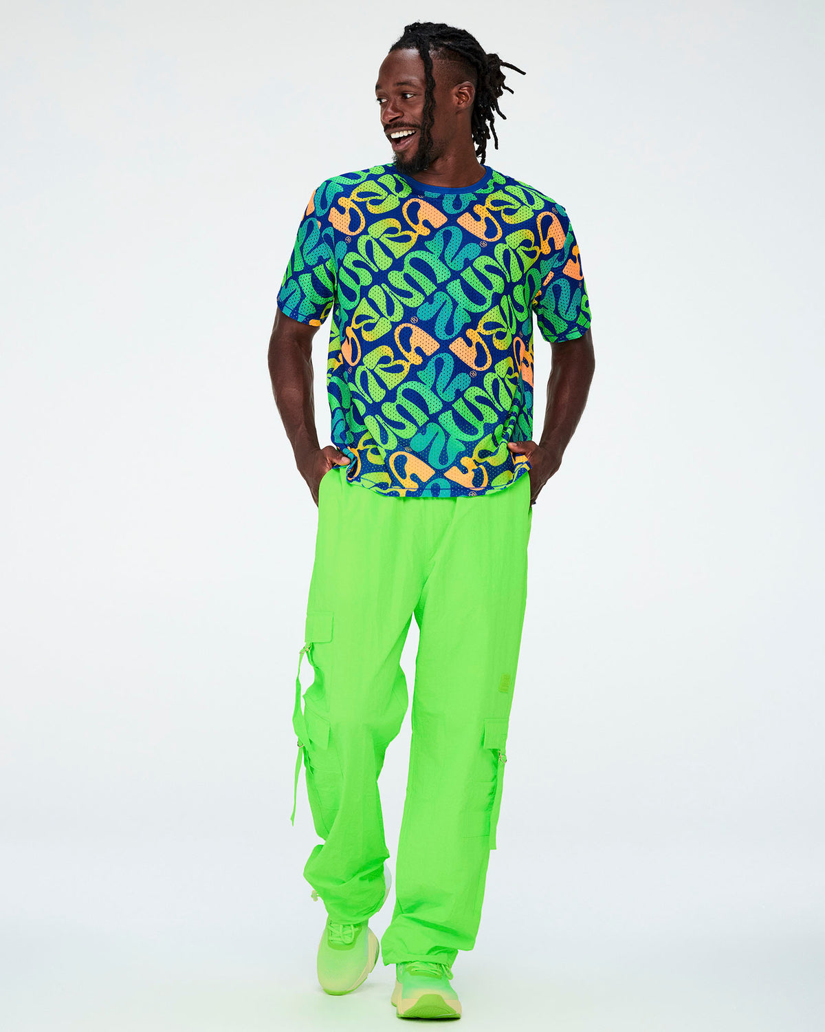 Full view of a male model in the blue warped 'ZUMBA' print mesh top with neon green cargo pants with dangling tassels and neon green shoes.