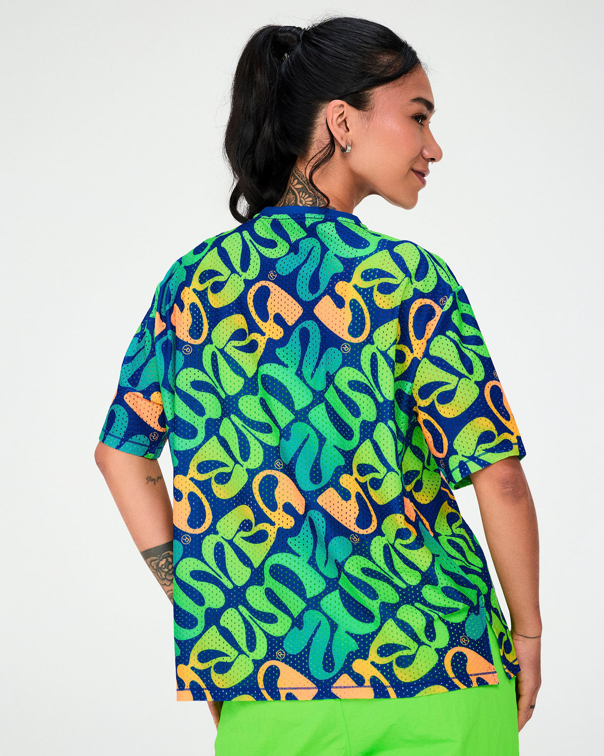 Back view of a female model in the blue warped 'ZUMBA' print mesh boxy top, which falls loosely to the hip over neon green pants.