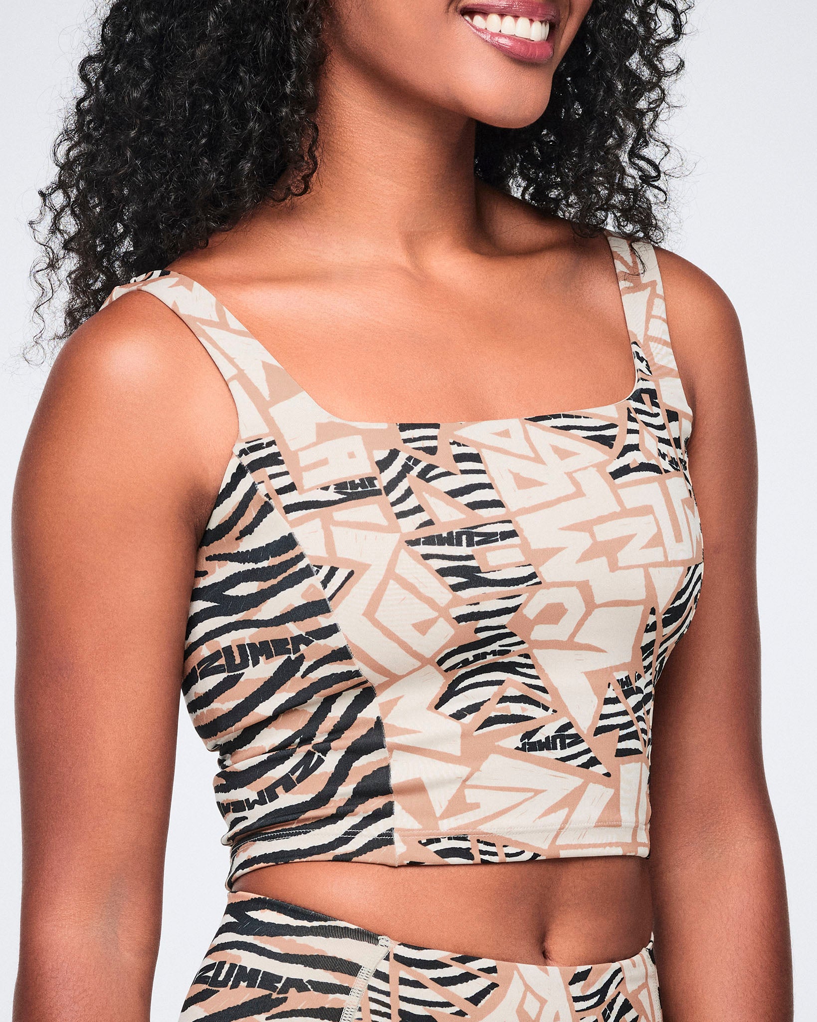 Female model wearing a fitted cropped tank in a beige, cream, and black abstract zebra-print pattern with a square neckline, wide shoulder straps, and a smooth body-hugging fit.