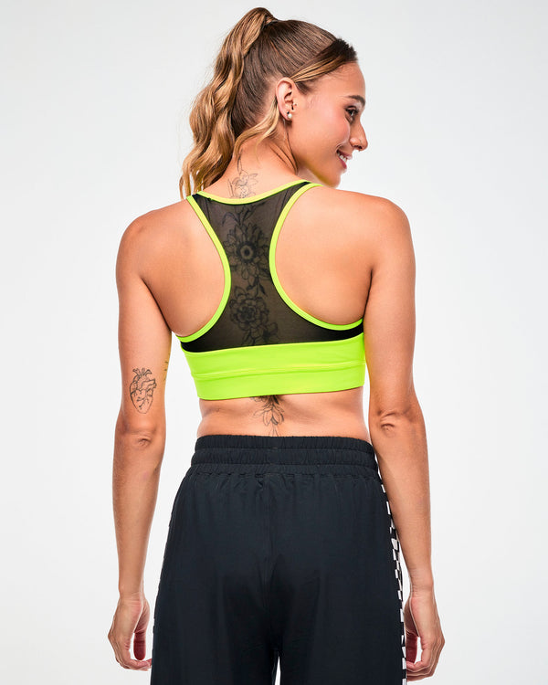 Back view of a female model in a yellow-green sports bra with a mesh racerback panel, paired with black pants.