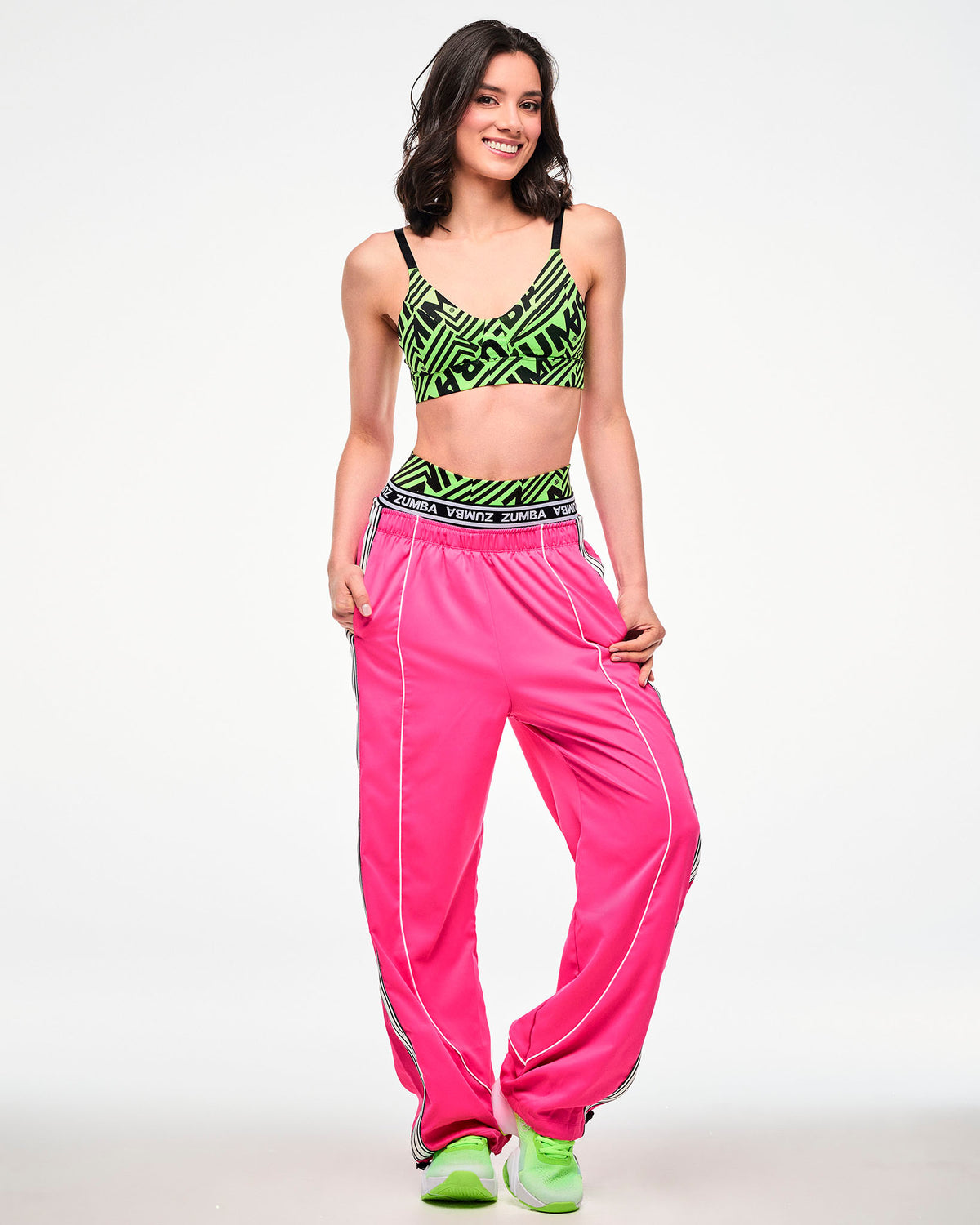 Full view of a female model wearing a neon green and black geometric striped print bra, paired with pink track pants and green shoes.