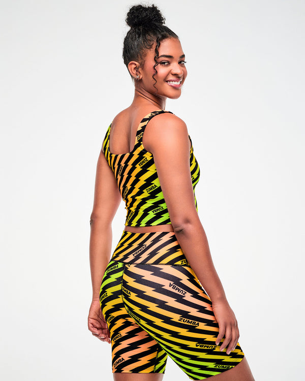 Female model wearing a yellow, green, and orange ombre crop top and matching biker shorts with a black lightning bolt zigzag print, side view.