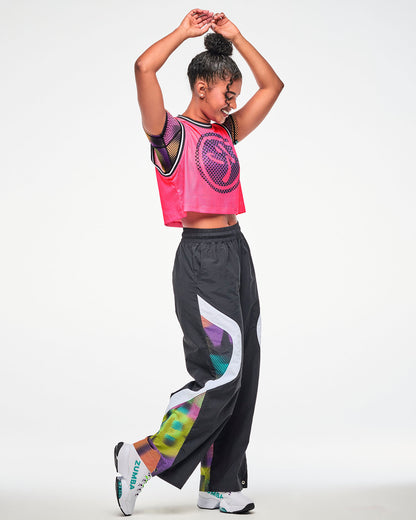 Female model wearing a pink mesh tank, paired with black wide-leg pants with a thick white strip and colorful Zumba print and white shoes.